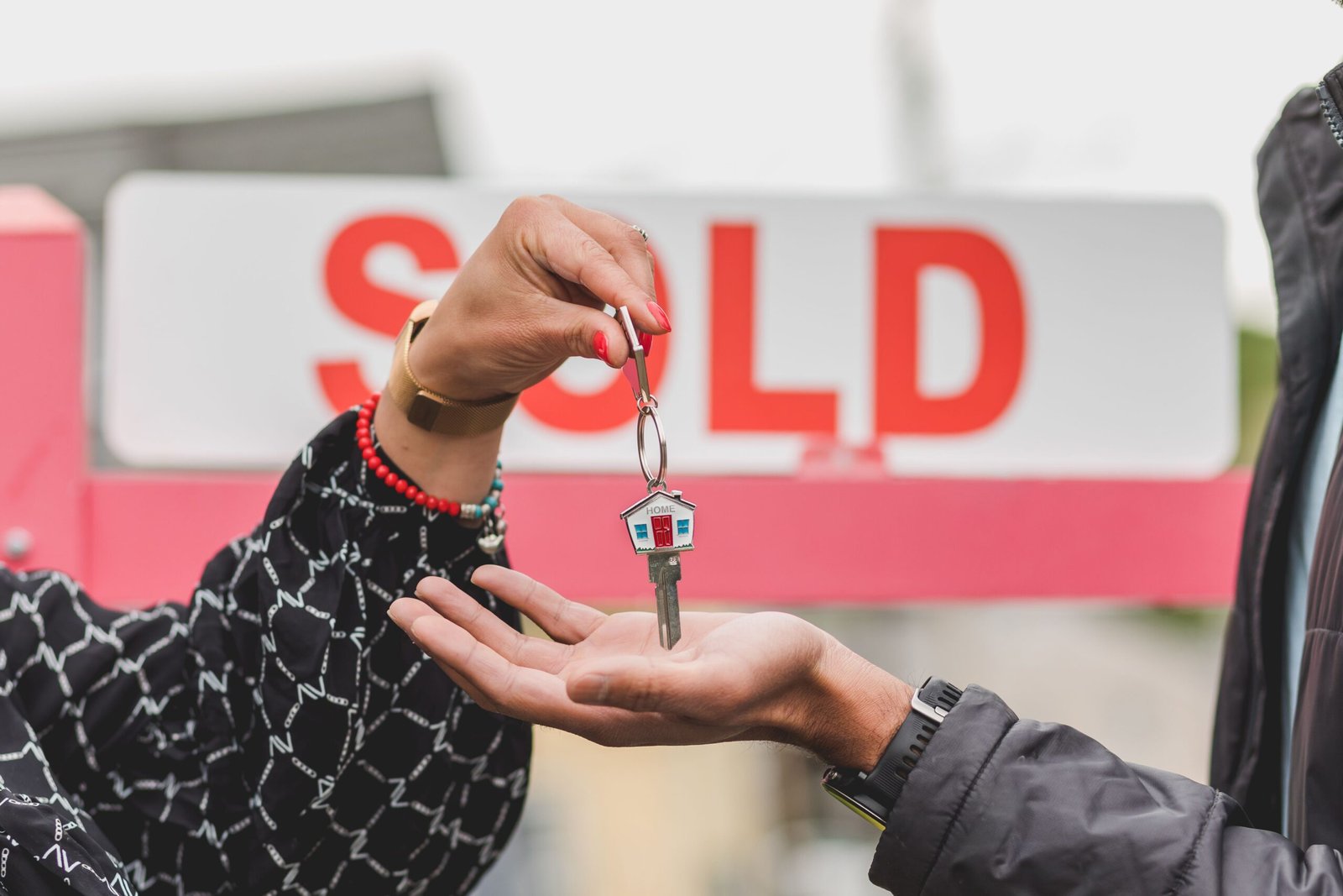 Should You Sell Your Riverside Home Now or Wait? Close-up of handing over house key with sold sign in background, symbolizing real estate success.